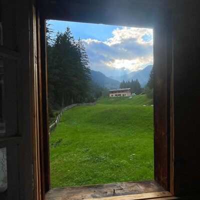 An idyllic view from the Alm's rustic wooden window overlooking lush green meadows and the surrounding mountain landscape.