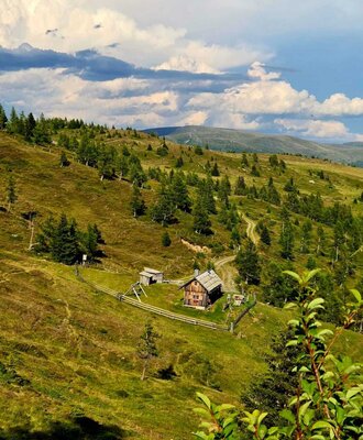 The rustic Alm nestled in a rolling, green mountain landscape under a vast cloudy sky.