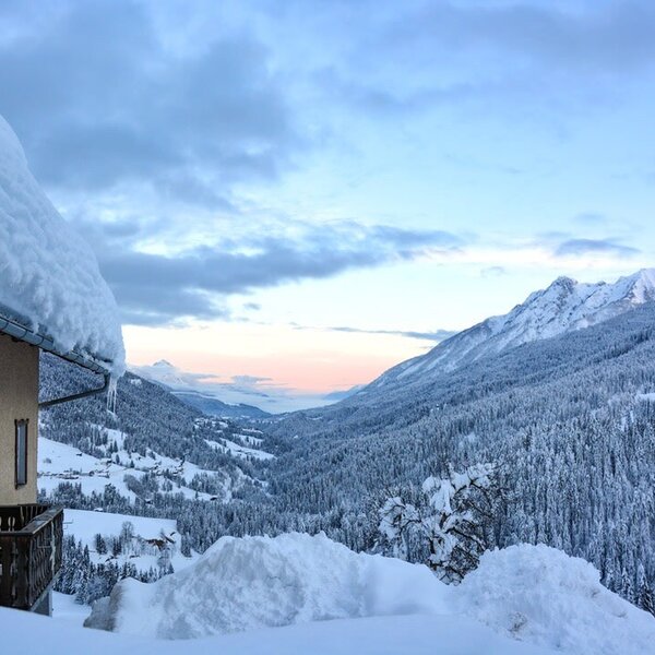 The farmhouse in winter, featuring a snow-covered roof, a balcony, and views of the surrounding mountain valley with forested slopes.