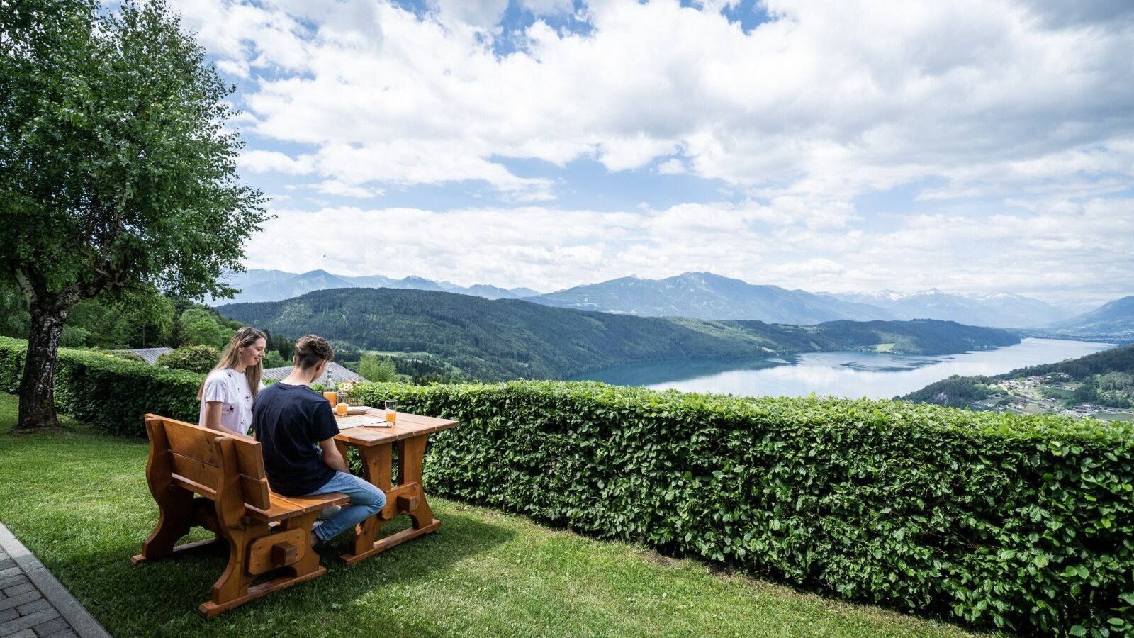 Outdoor seating area at the Ferienhaus with a wooden table and bench, offering panoramic views of the lake and mountains.