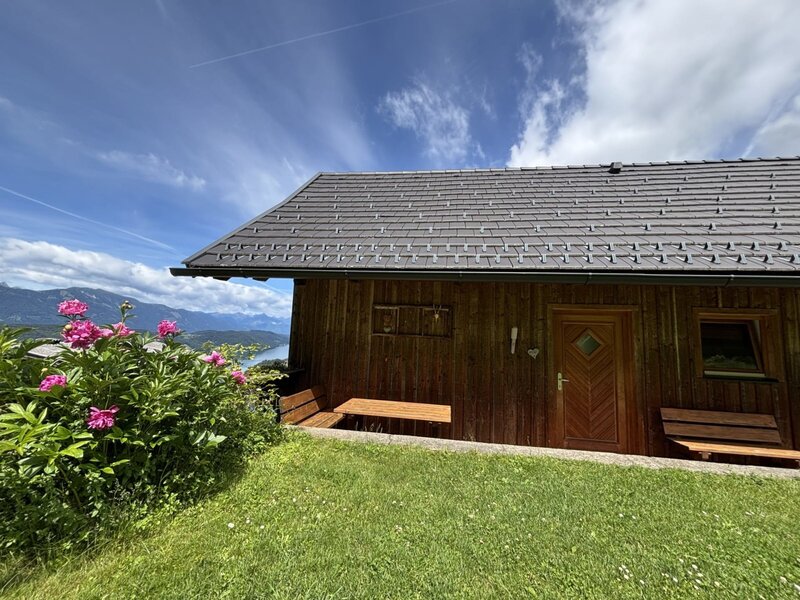 Exterior of the Ferienhaus featuring a wooden facade, roof, and outdoor benches on a lawn, with peonies and a view of the lake and mountains.