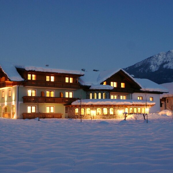 The farm house exterior at night, with illuminated windows and snow-covered ground, set against snowy mountains.