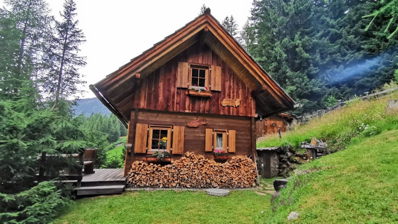 Exterior view of the Alm with a wooden facade, window shutters, flower boxes, a stack of firewood, and a small wooden terrace.