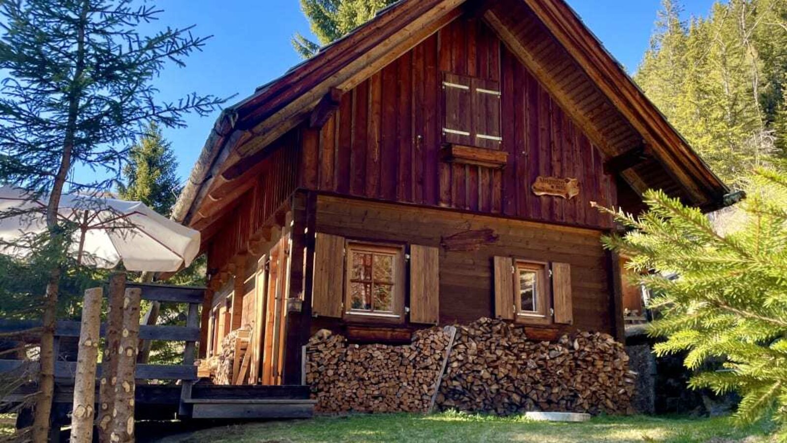 The Alm exterior, featuring a wooden facade, gabled roof, shutters, and a stack of firewood in front of a grassy area.