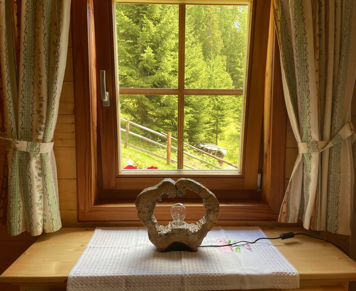 An inviting view of the lush green forest from the Alm's wooden window, featuring a rustic tabletop lamp.