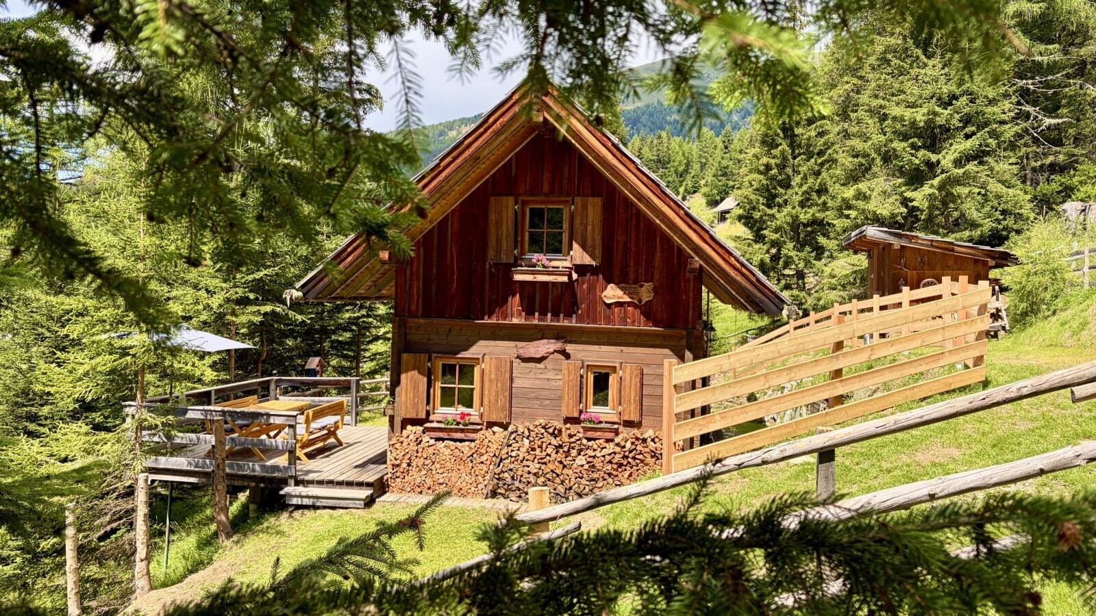 A rustic Alm featuring an inviting wooden terrace, stacked firewood, and lush forest surroundings.