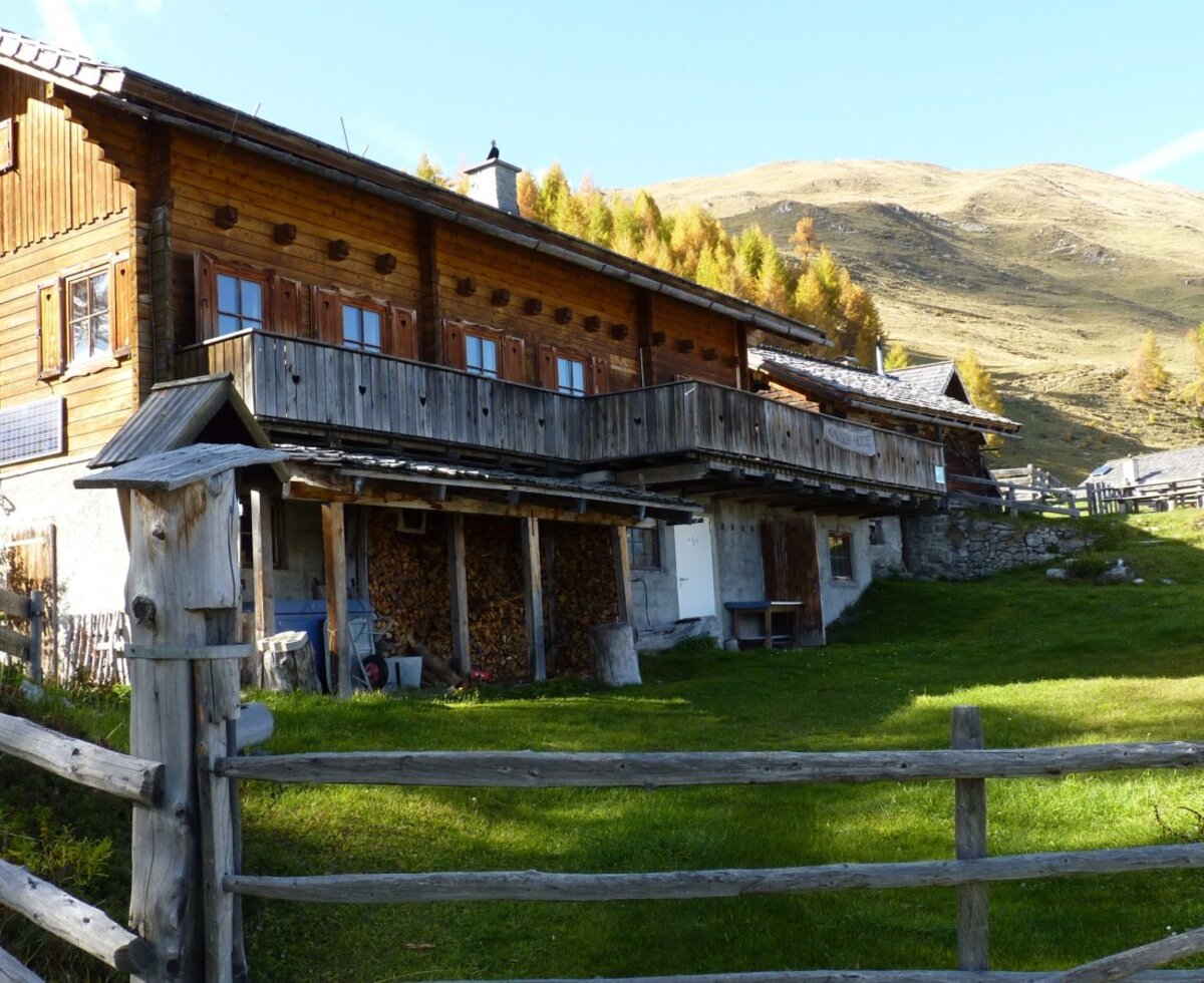 Exterior of the wooden Alpine Hut, featuring balconies, window shutters, a solar panel, and stacked firewood, set within a fenced grassy area with a mountain backdrop.