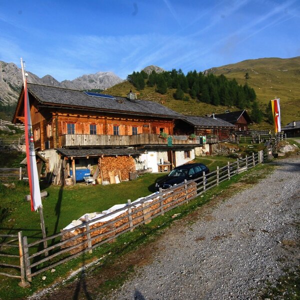 The main wooden Alpine Hut building, featuring a balcony, stacked firewood, and surrounding mountain landscape, accessible via a gravel path.
