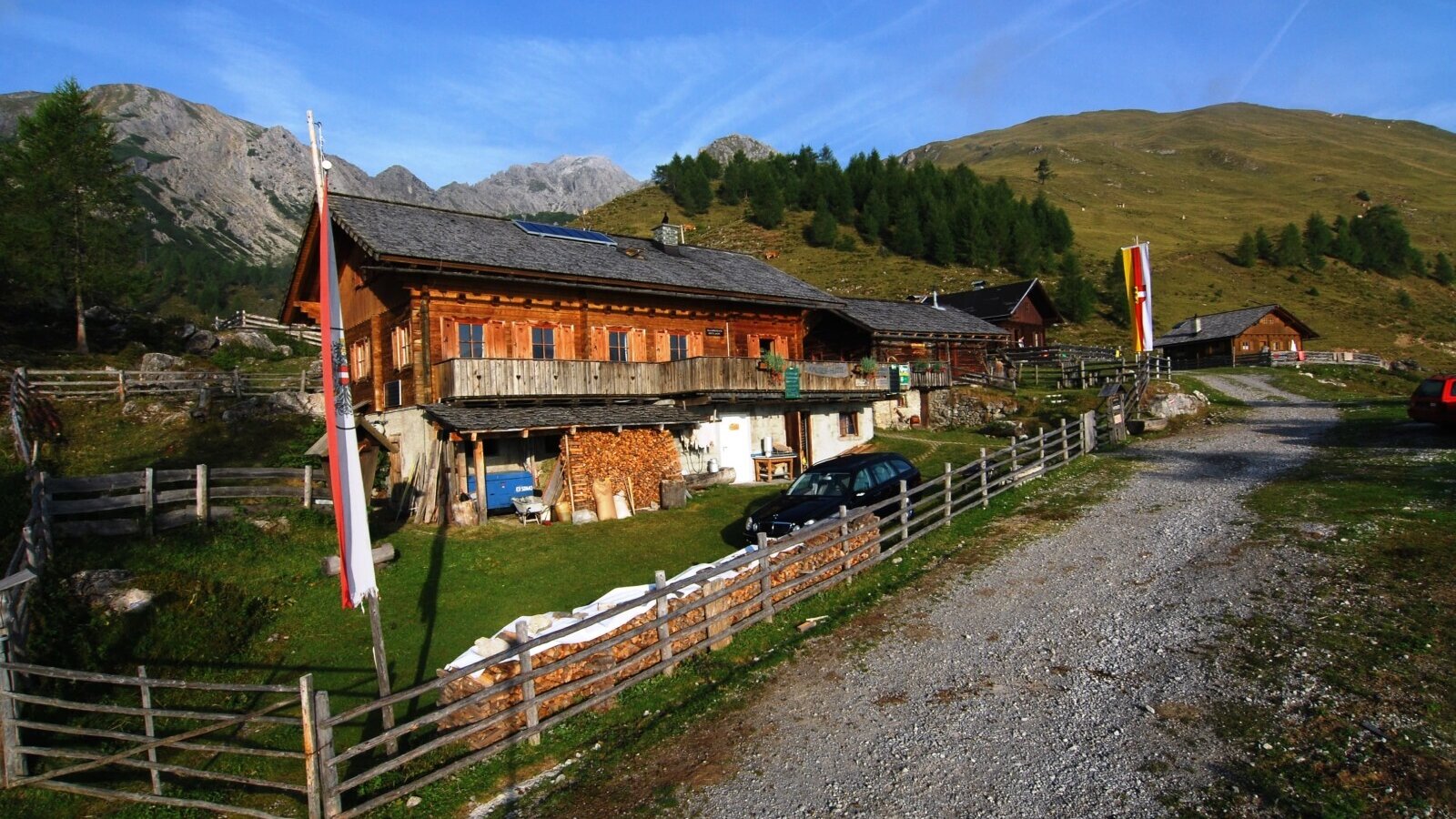 The main wooden Alpine Hut building, featuring a balcony, stacked firewood, and surrounding mountain landscape, accessible via a gravel path.