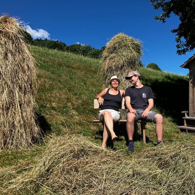 Guests on a wooden bench among haystacks, with a 'Milch-Mobil' shed, showcasing the rural setting of the Bauernhof.