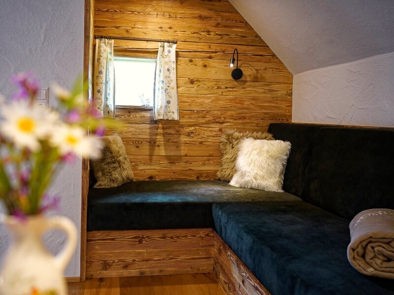 Corner seating area within the Farm House, featuring wooden paneling, dark cushions, furry pillows, and a window.
