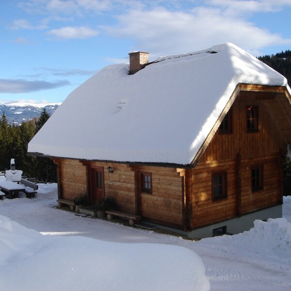 The wooden Alpine Hut, featuring a snow-covered roof and exterior, set in a winter landscape with mountain views.