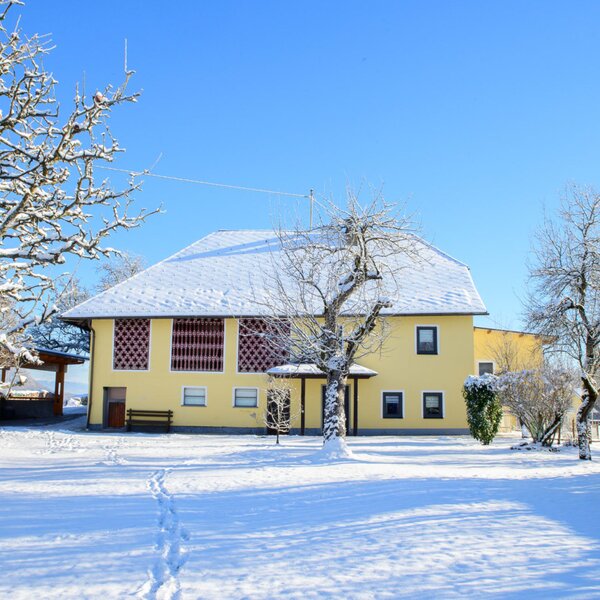 Exterior view of the yellow farmhouse with a snow-covered roof and winter landscape under a blue sky.