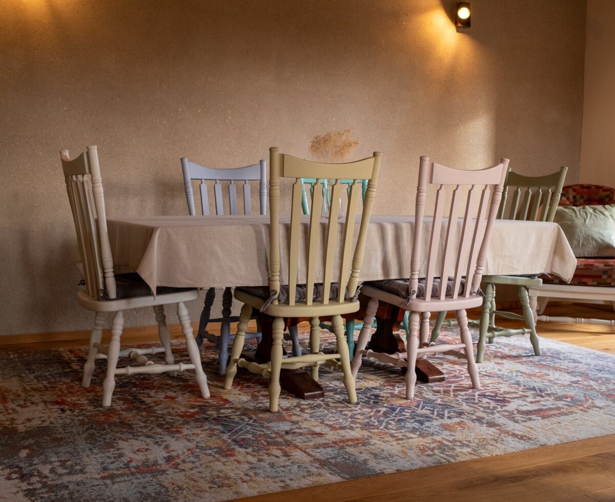 The farm house dining area features a table with a tablecloth, several chairs in varied pastel colors, and a patterned rug on a wooden floor.