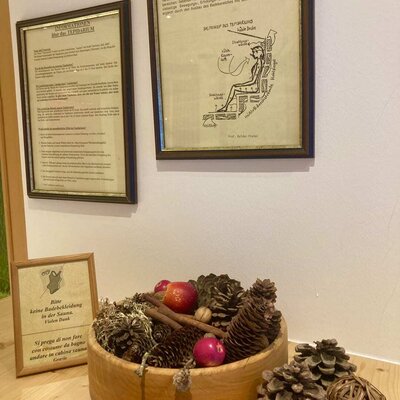 A decorative wooden bowl filled with pine cones, cinnamon sticks, and apples, displayed alongside framed information about the Tepidarium and sauna rules at the Bauernhof.