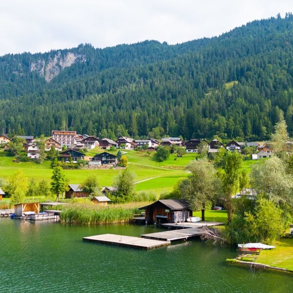 A village on the shore of Lake Weissensee with forested mountains in the background, showing the surroundings of the farmhouse.