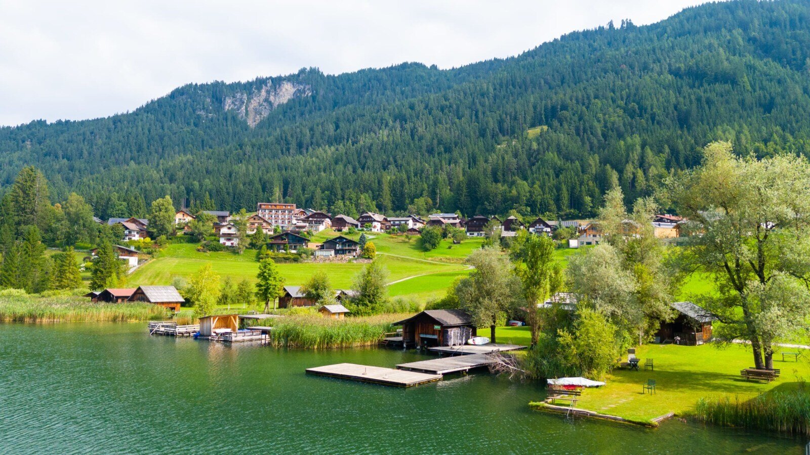 A village on the shore of Lake Weissensee with forested mountains in the background, showing the surroundings of the farmhouse.