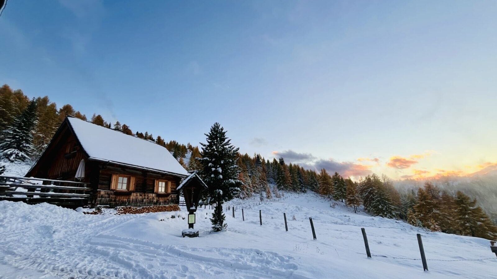 The snow-covered Alm with lit windows and stacked firewood, surrounded by a winter landscape.
