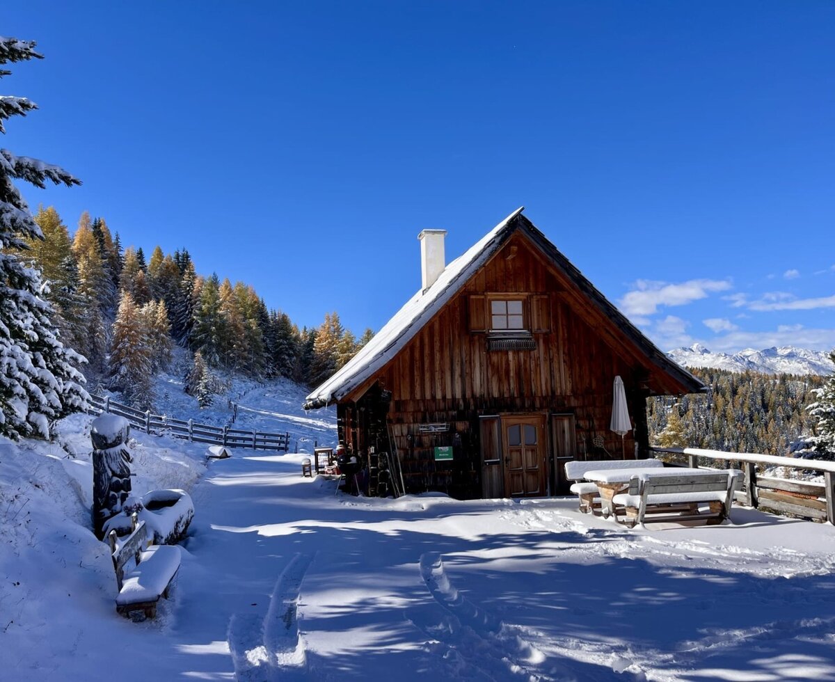 The alm in winter, surrounded by snow and mountains, with a snowy wooden table and benches outside.