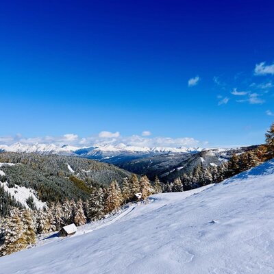 Winter landscape with snow-covered mountains, forests, and Alm huts under a blue sky.