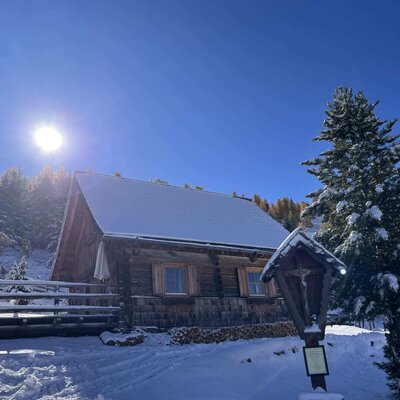 The snow-covered Alm under a blue sky on a sunny winter day, with a wayside shrine in the foreground.