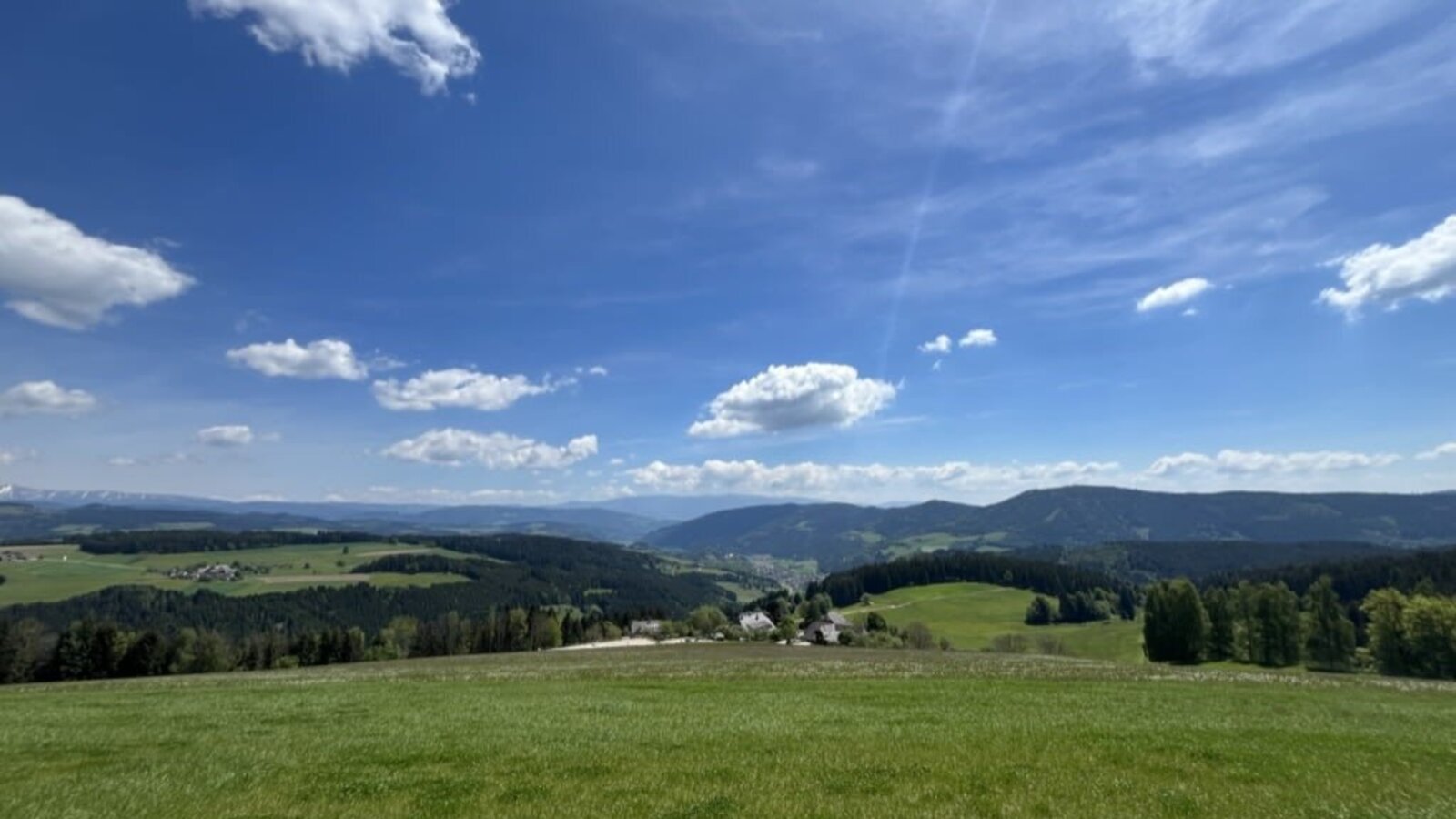 Expansive view from the farmhouse over green fields, forests, and the surrounding hilly landscape under a blue sky.