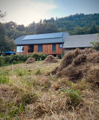 The farmhouse featuring a wooden facade and solar panels on the roof, set on a hillside with a forest in the background.
