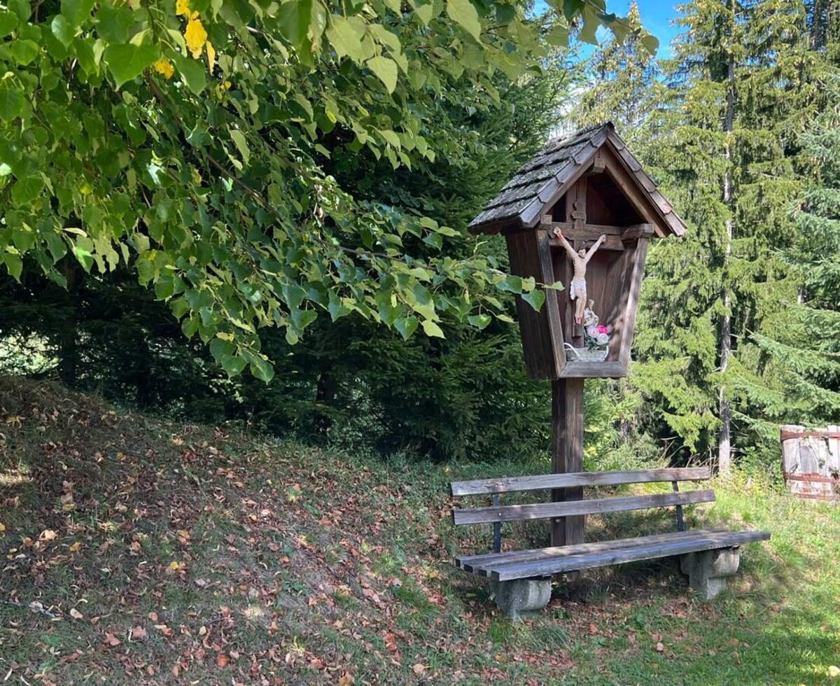 A peaceful resting spot at the farmhouse with a rustic wooden bench and a traditional wayside cross by the forest edge.