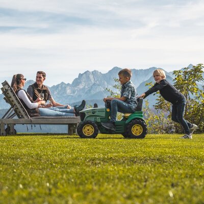 The farmhouse features an outdoor area with a lounge chair and a children's toy tractor, set against mountain views.