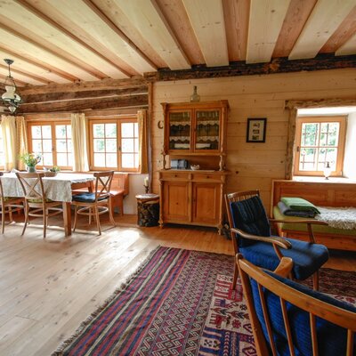 The farmhouse features a spacious living and dining area with a wooden beamed ceiling, a dining table with bench seating by large windows, and a separate seating area with wooden armchairs and a daybed.