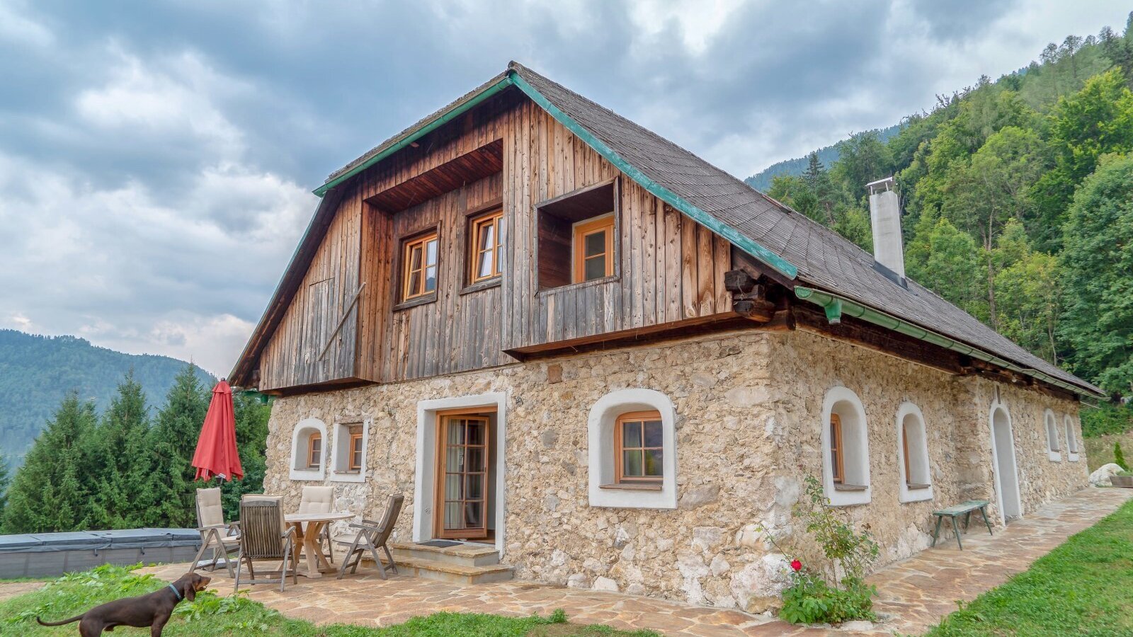 The Alpine Hut features a stone ground floor and a wooden upper level, with an outdoor patio equipped with a dining table, chairs, and a red umbrella.