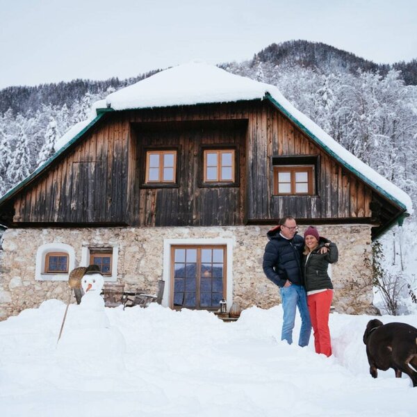 The Alpine Hut exterior, featuring a stone ground floor and wooden upper levels, is surrounded by snow with a couple, a dog, and a snowman in the foreground.