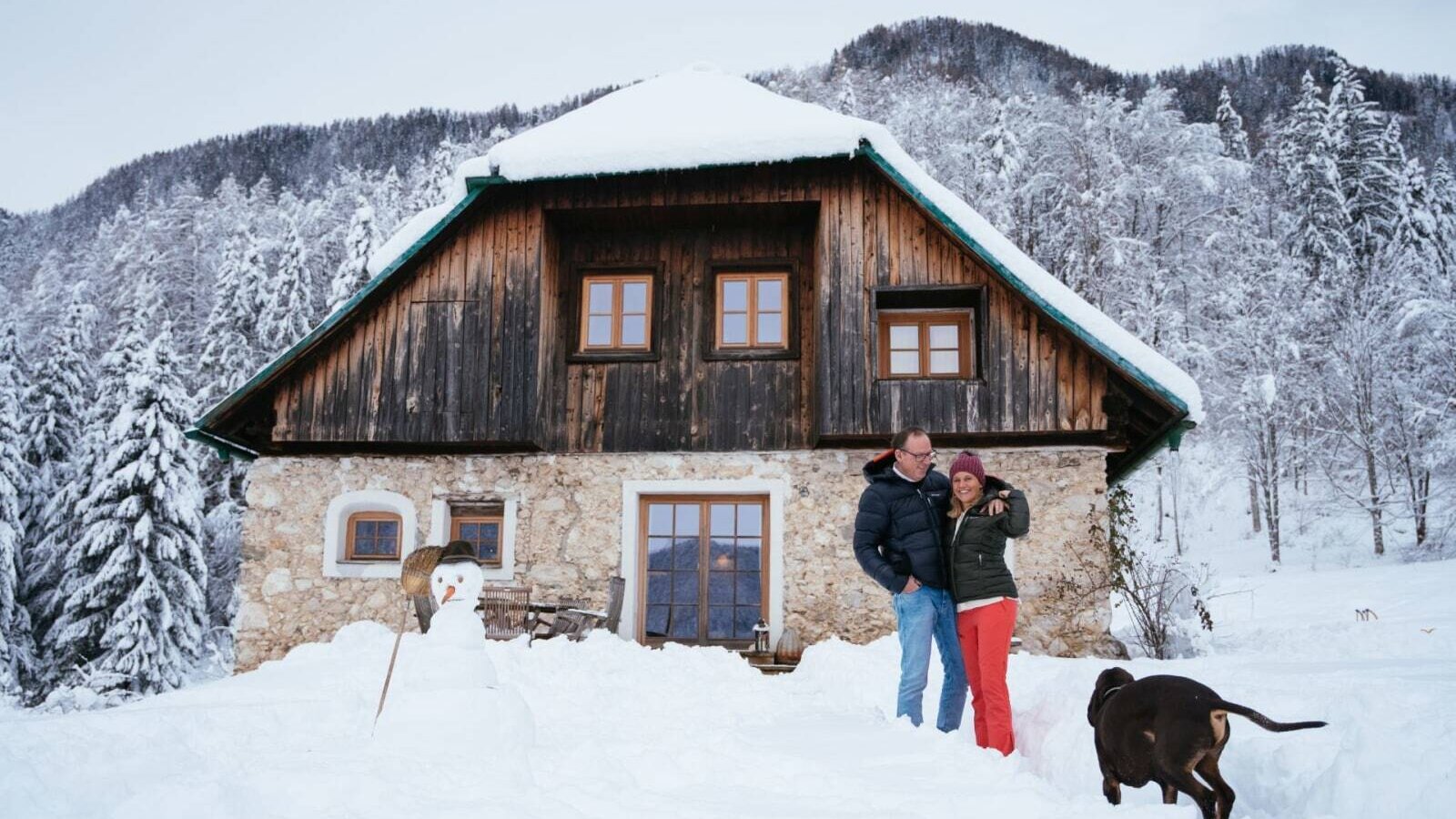 The Alpine Hut exterior, featuring a stone ground floor and wooden upper levels, is surrounded by snow with a couple, a dog, and a snowman in the foreground.