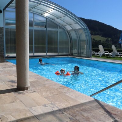 The partially covered swimming pool of the Ap­par­te­ments with a sunbathing lawn and mountain view.