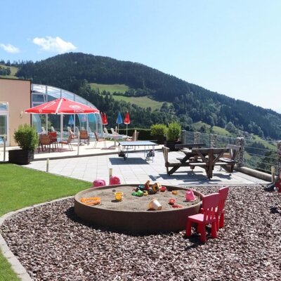 Outdoor area of the Ap­par­te­ments with a lawn, children's playground, terrace with seating, table tennis table, pool area, and mountain views.