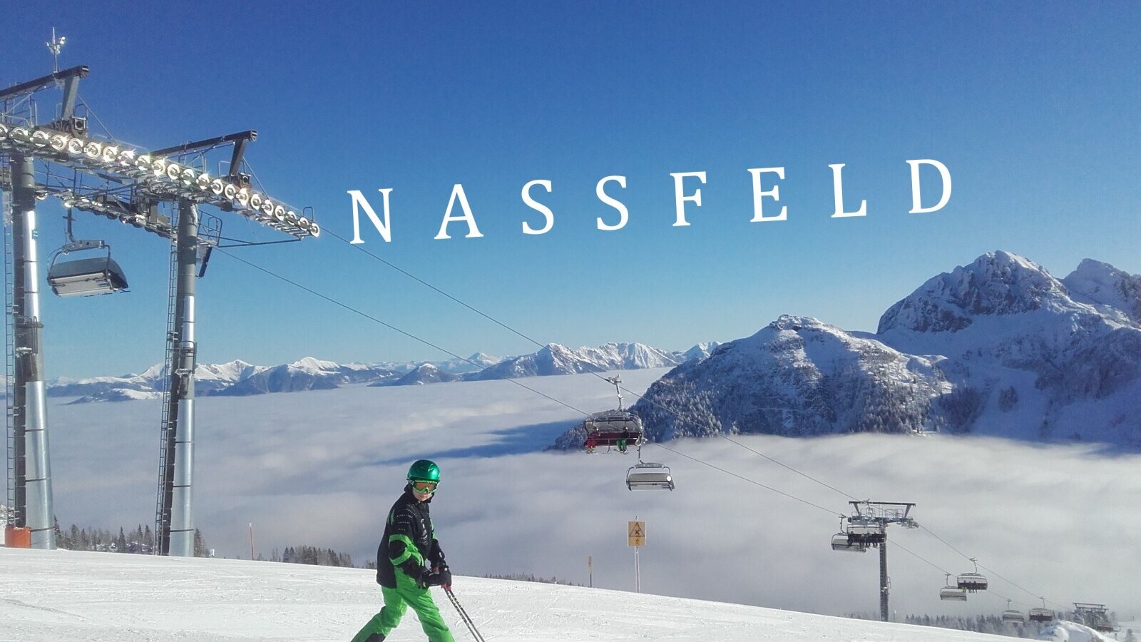 A skier on the Nassfeld slopes, with ski lifts, snow-capped mountains, and clouds filling the valleys below.