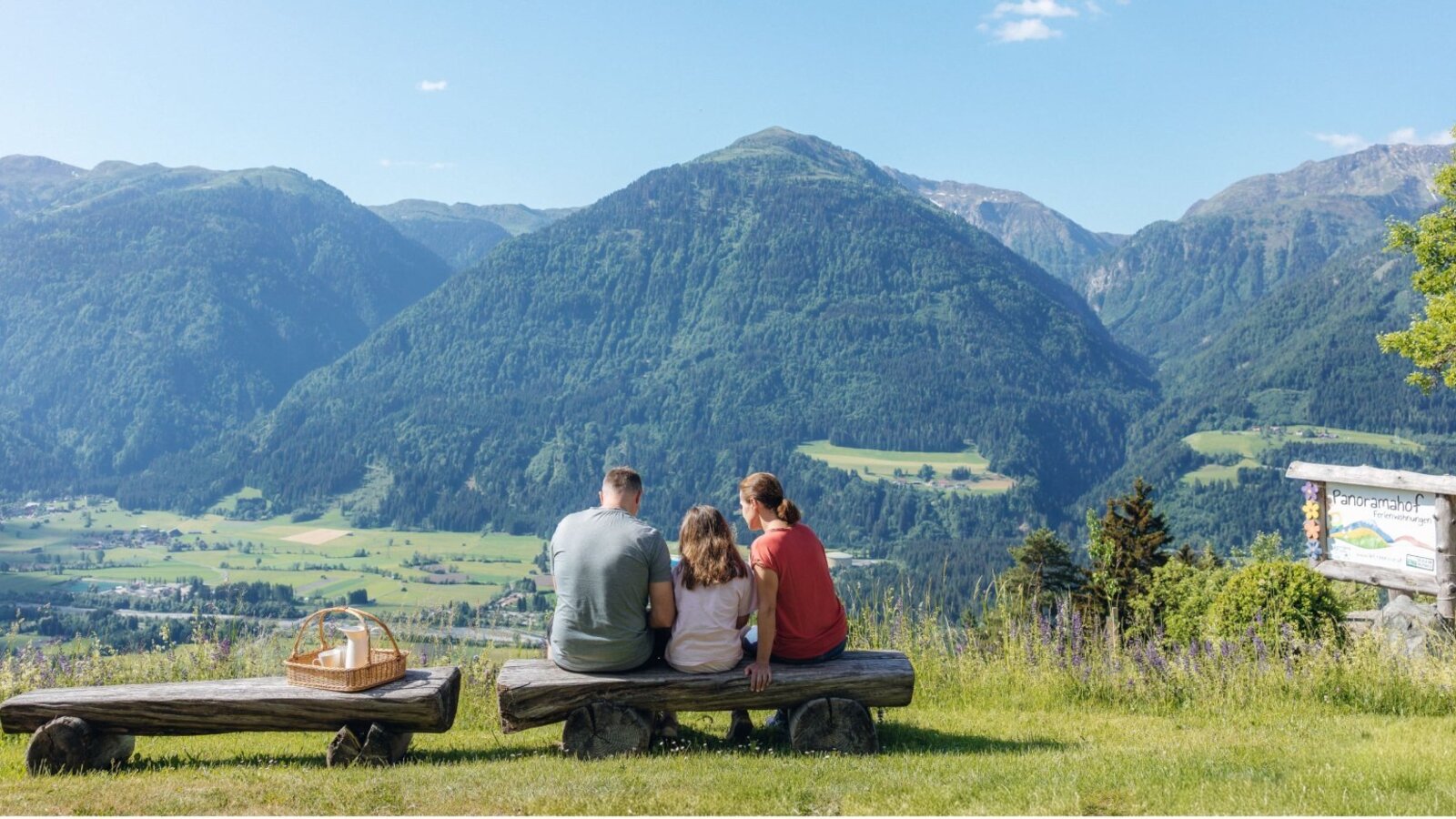 A family enjoys the panoramic view of the mountains and valley from the Panoramahof farmhouse, seated on wooden benches with a picnic basket.