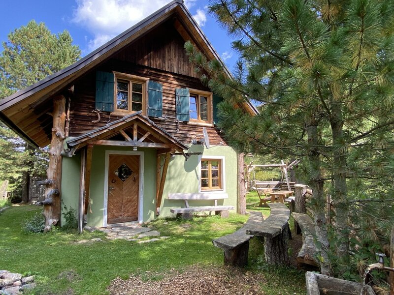 The exterior of the Alpine Hut, featuring a wooden facade, green shutters, and an outdoor seating area with carved wooden benches.