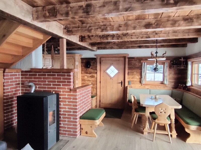 The Alpine Hut's living and dining area features a wood-burning stove with a brick base, a corner dining bench with wooden furniture, and stairs leading to the upper level.