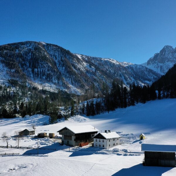 The farmhouse complex in a snow-covered valley, surrounded by forested mountains under a clear sky.