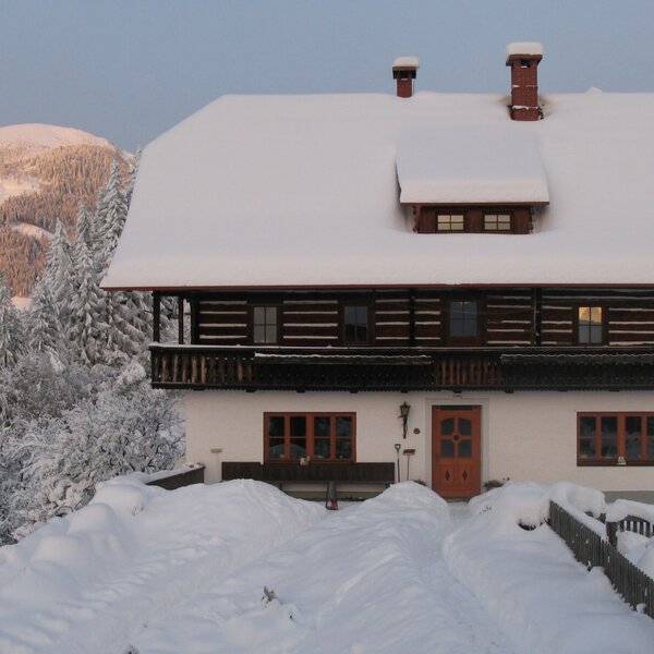 The snow-covered farm house features a wooden balcony and is surrounded by snowy mountains and trees.