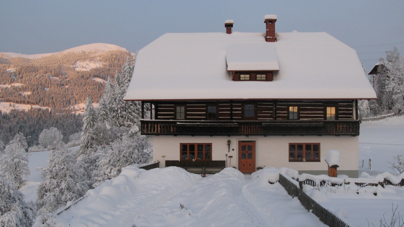 The snow-covered farm house features a wooden balcony and is surrounded by snowy mountains and trees.