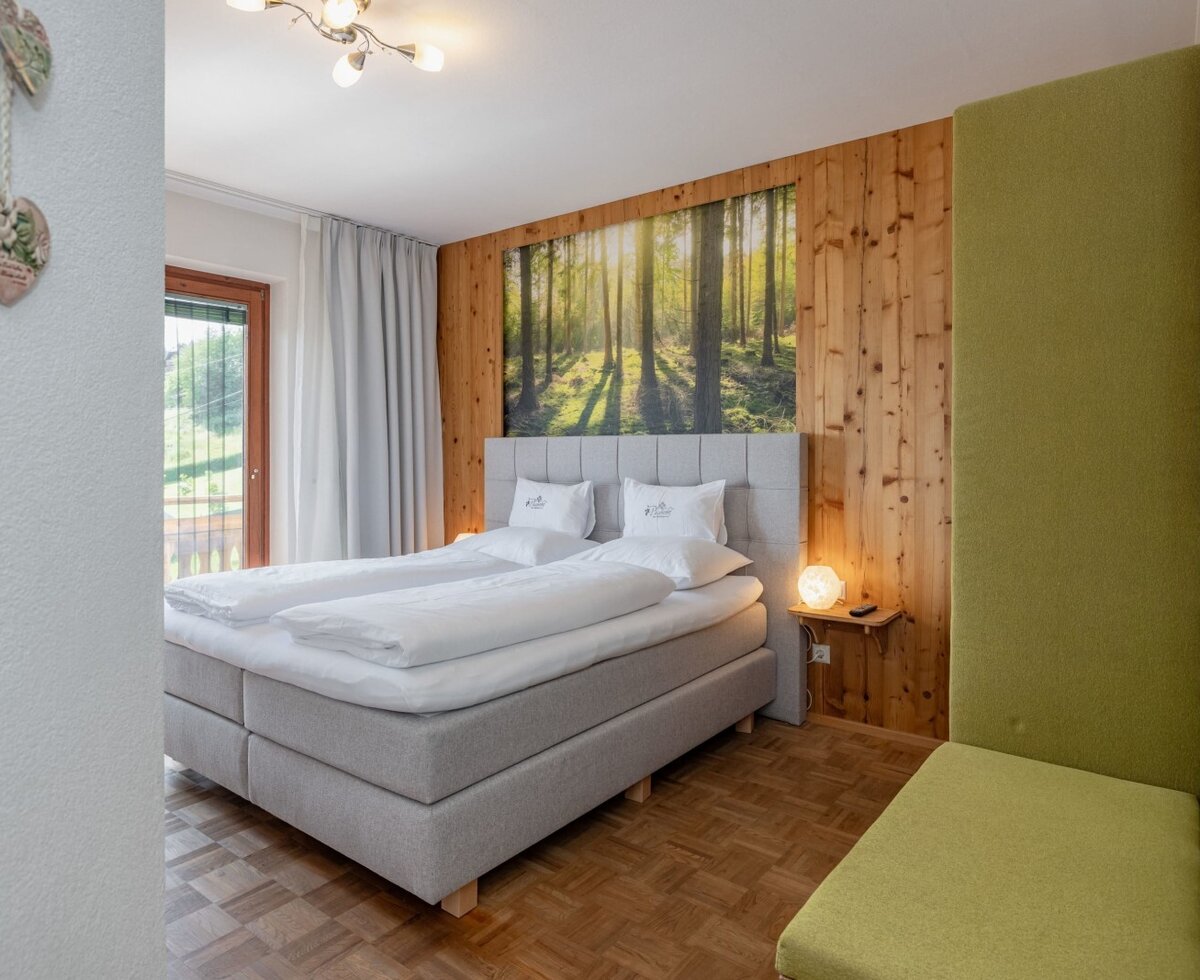 The Farm House bedroom features a double bed, wooden wall paneling with a forest print, a green cushioned bench, and balcony access.
