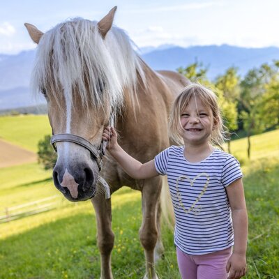 A young girl stands next to a horse in a green field at the Farm House, with mountains visible in the background.