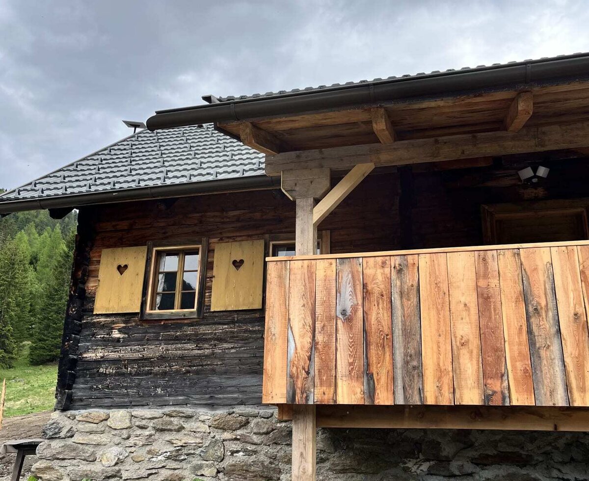 A traditional wooden Alm featuring a rustic balcony, solid stone foundation, and charming window shutters with heart cut-outs.