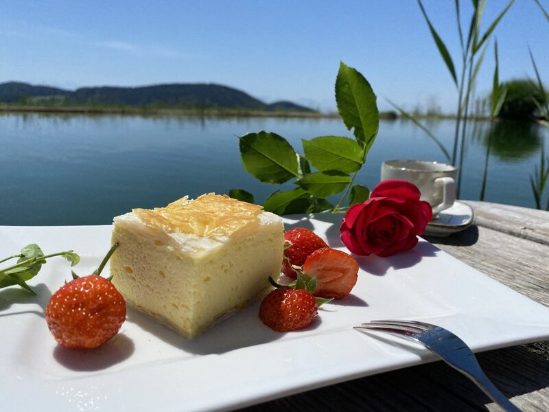 A slice of cream cake with strawberries and a cup of coffee, served on a wooden table beside the water at the Farm House.