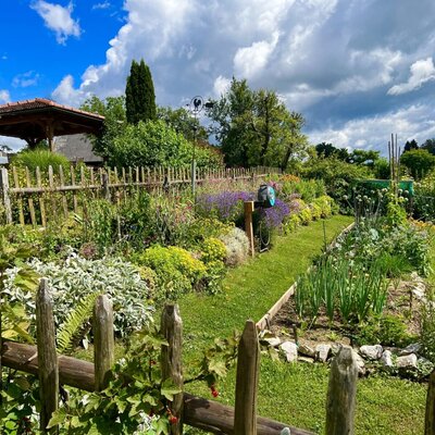 The garden of the farmhouse featuring a wooden fence, diverse flower beds, and a vegetable patch.