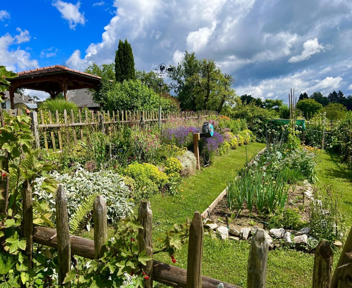 The garden of the farmhouse featuring a wooden fence, diverse flower beds, and a vegetable patch.
