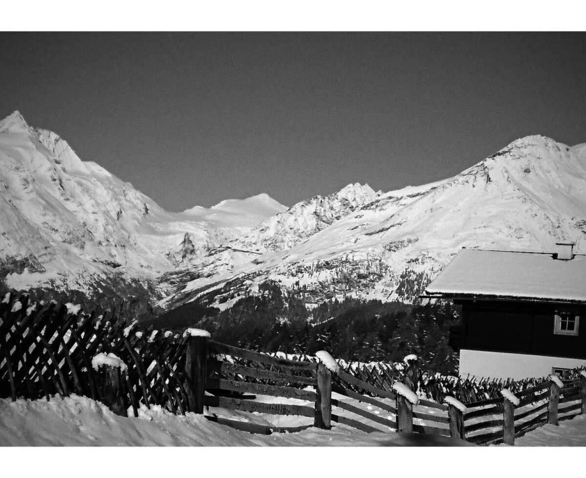 The Alpine Hut in a snow-covered mountain landscape, featuring a traditional wooden fence and panoramic views of the surrounding peaks.