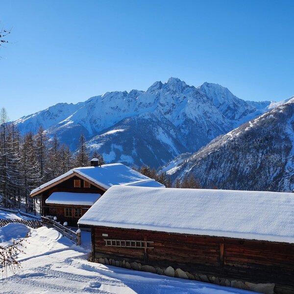 The snow-covered Almhütte and a second wooden building, surrounded by a winter landscape with snow-capped mountains.